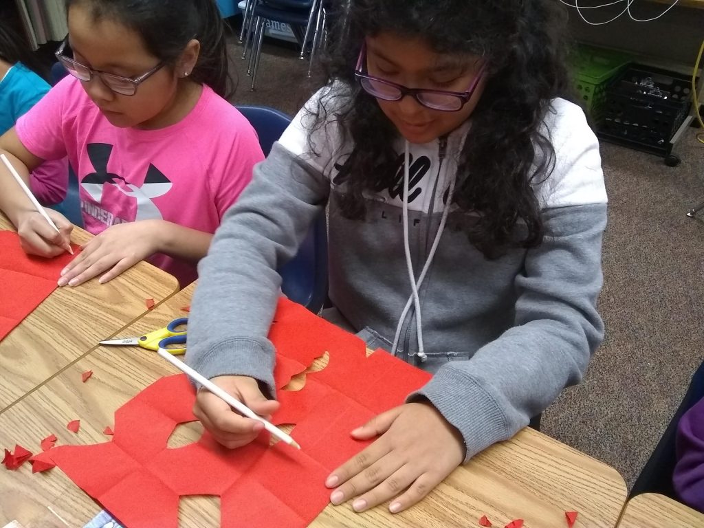 Two Latina youth sit at desks, drawing with white pencils a shape to cut out of their red paper.