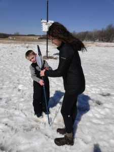 Young boy stands in the snow while teaching a female St. Olaf student how to hold an atlatl.