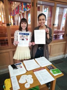 Two female St. Olaf students stand holding materials in front of a table outside of the chapel.