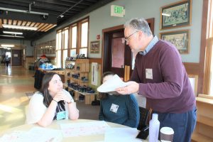 Two female students sit at round table while Dale Kruse, opera creation instructor, stands and gives them directions.