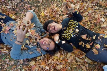A fall scene of two student playing with fallen leaves on the campus greens.