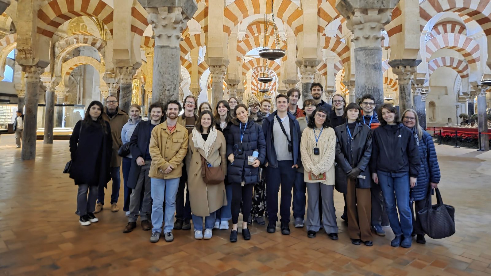 A large group of young adults and a few older individuals posing for a photo inside the Mosque-Cathedral of Córdoba, Spain, featuring the iconic red-and-white double arches and stone columns.
