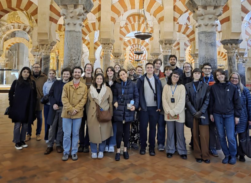 A large group of young adults and a few older individuals posing for a photo inside the Mosque-Cathedral of Córdoba, Spain, featuring the iconic red-and-white double arches and stone columns.