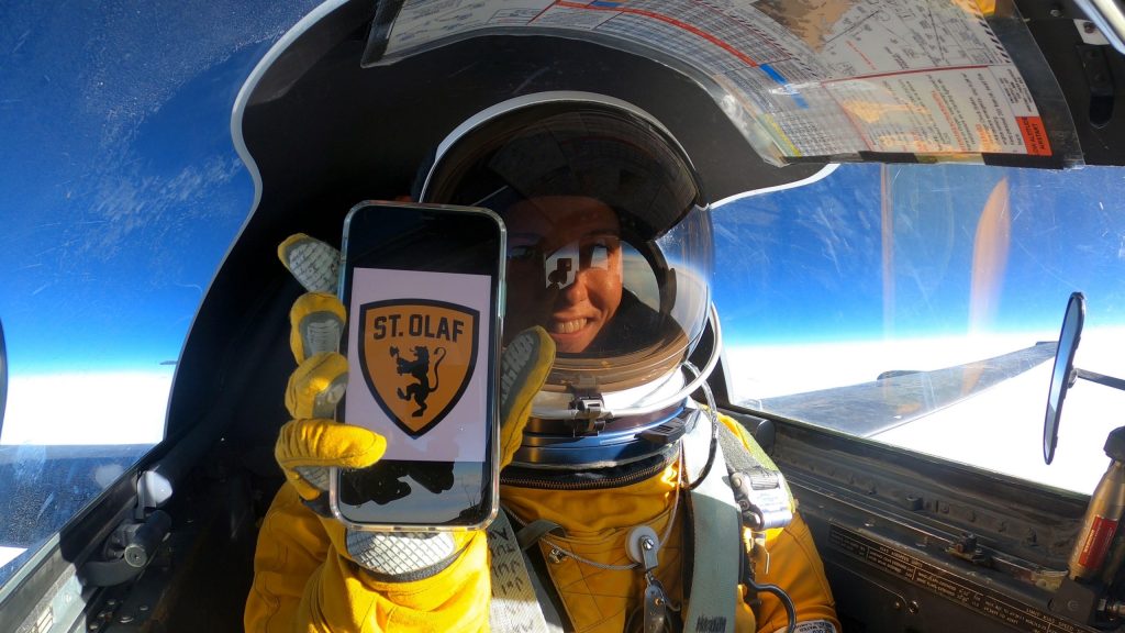 Michelle Cunningham, in a jet, holding a phone with the St. Olaf logo displayed.