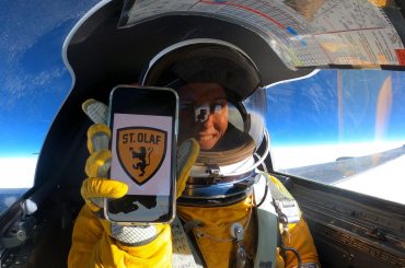 Michelle Cunningham, in a jet, holding a phone displaying the St. Olaf shield