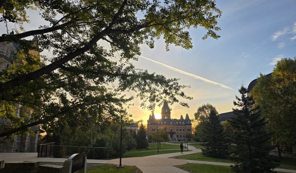 Image of Old Main in the distance with Holland Hall on the edge of the frame to the left. Dawn of day lighting. Trees framing the image on left and right.