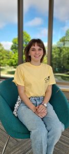 Katelyn sitting on a green chair, looking into the camera and smiling with short brown hair, wearing a yellow t shirt and jeans.
