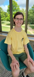 Max, seated on a green chair, with brown hair a little past his ears tucked behind his ears, a yellow shirt, and green shorts.  Max is looking into the camera and smiling
