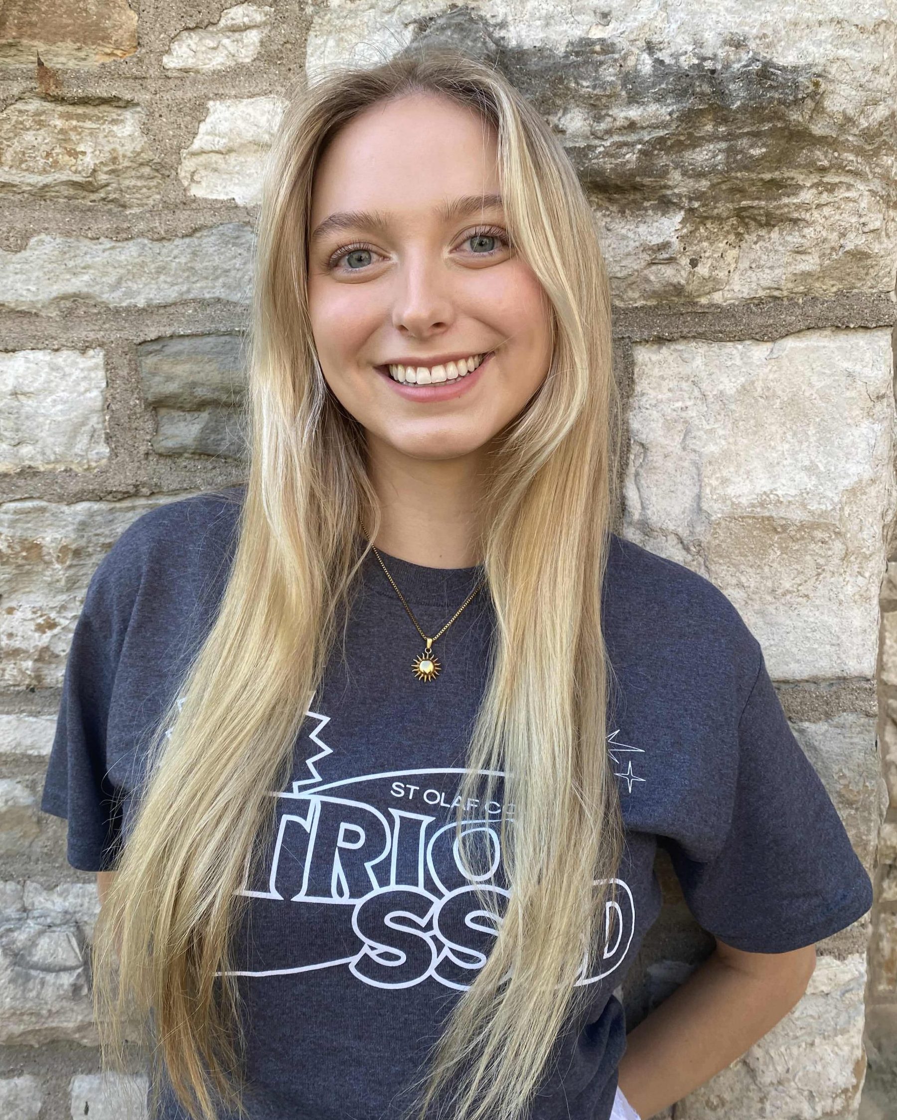 A smiling woman with long blonde hair and white skin. She is wearing the 2024 TRIO SSSD t-shirt and standing in front of a limestone brick wall.