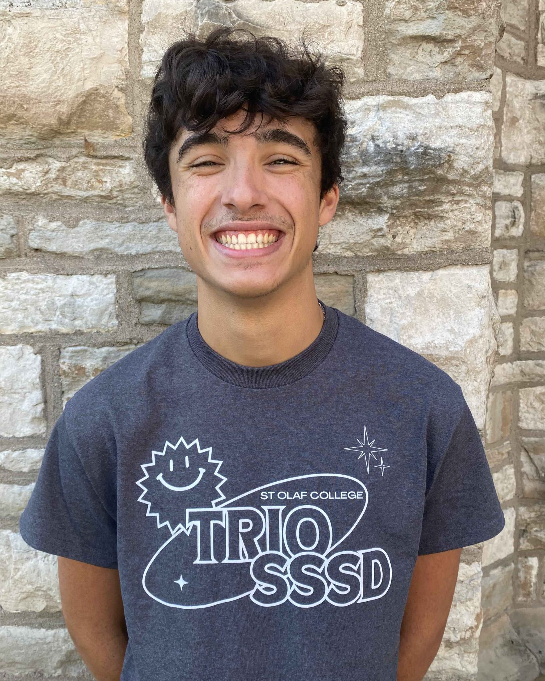 A smiling man with short brown hair and light brown skin. He is wearing the 2024 TRIO SSSD t-shirt and standing in front of a limestone brick wall