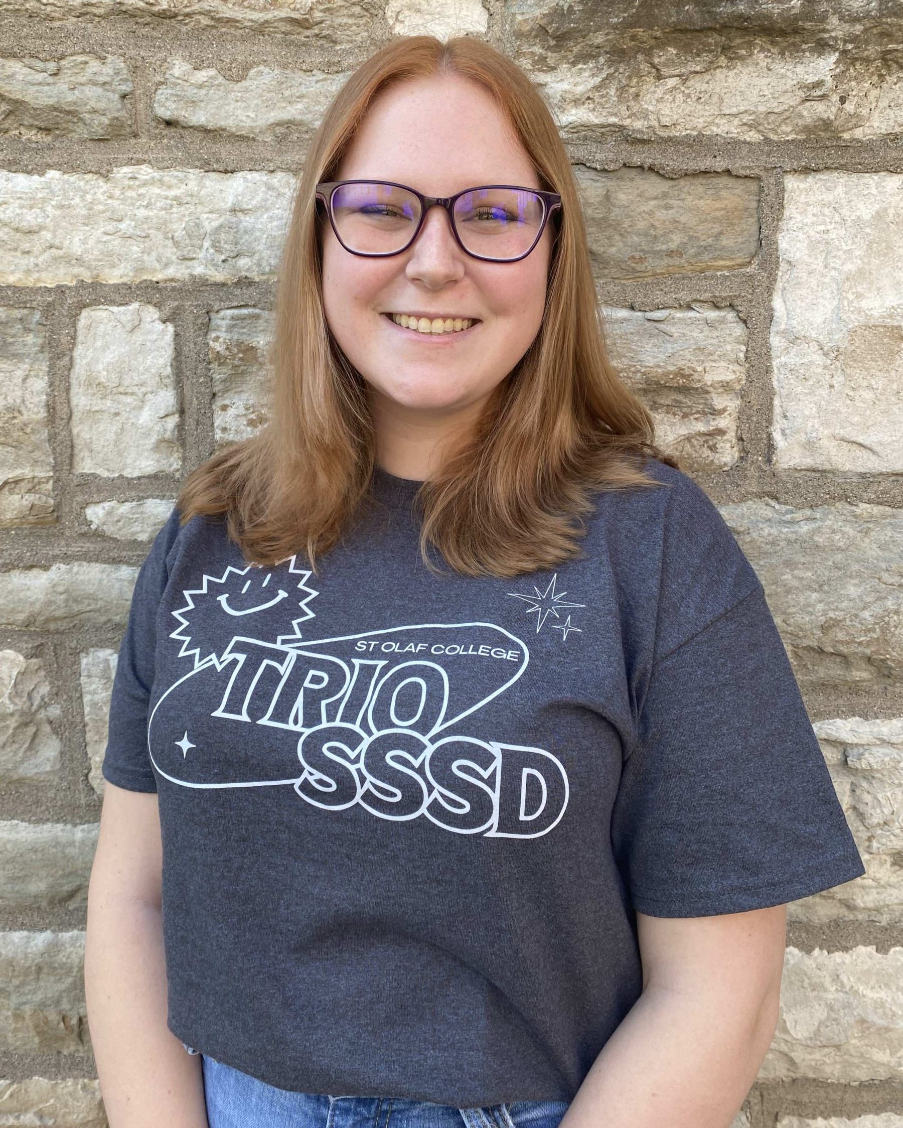 A smiling woman with glasses, medium length reddish-brown hair and fair white skin. She is wearing the 2024 TRIO SSSD t-shirt and standing in front of a limestone brick wall