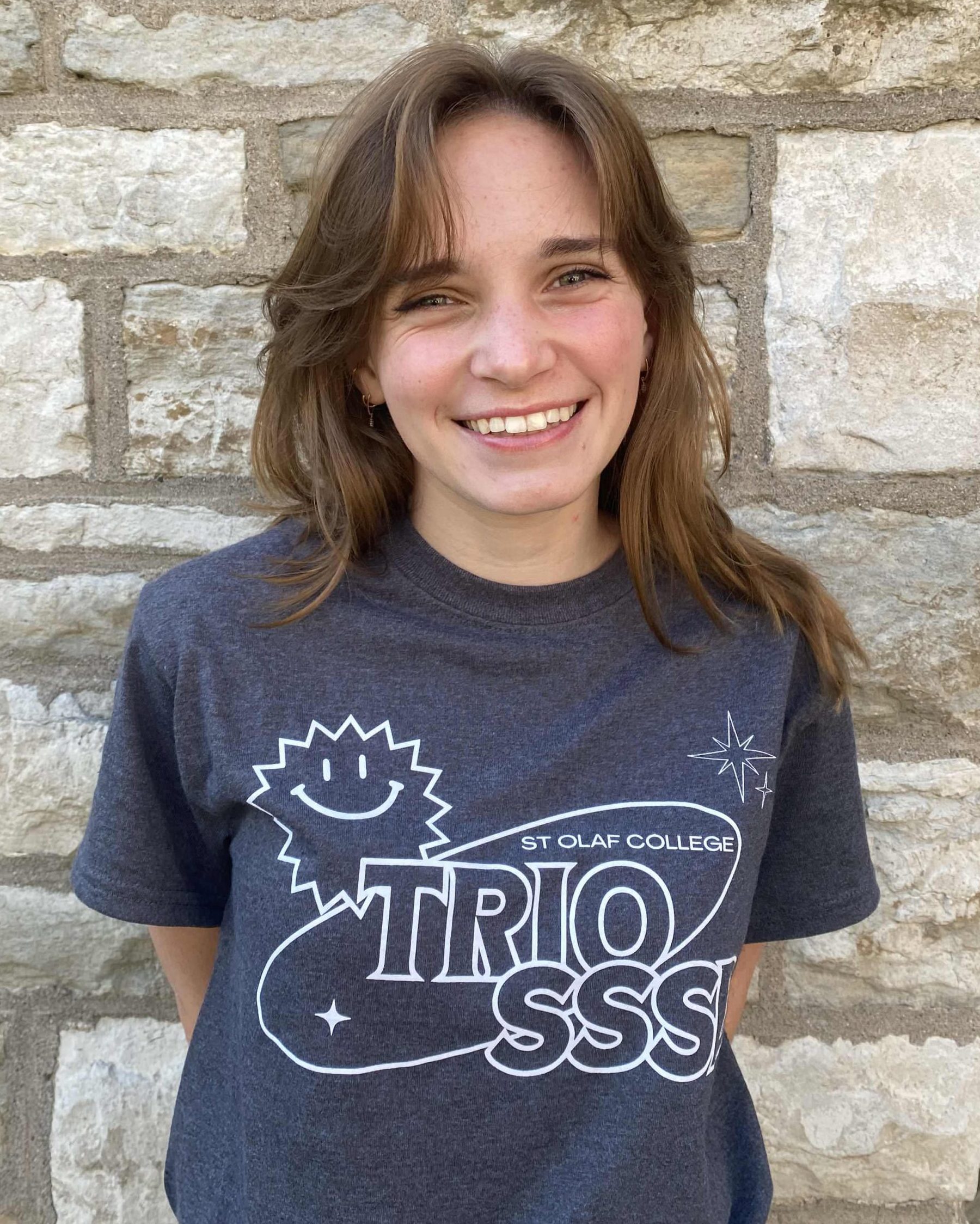 A smiling woman with medium length brown hair and white skin. She is wearing the 2024 TRIO SSSD t-shirt and standing in front of a limestone brick wall.