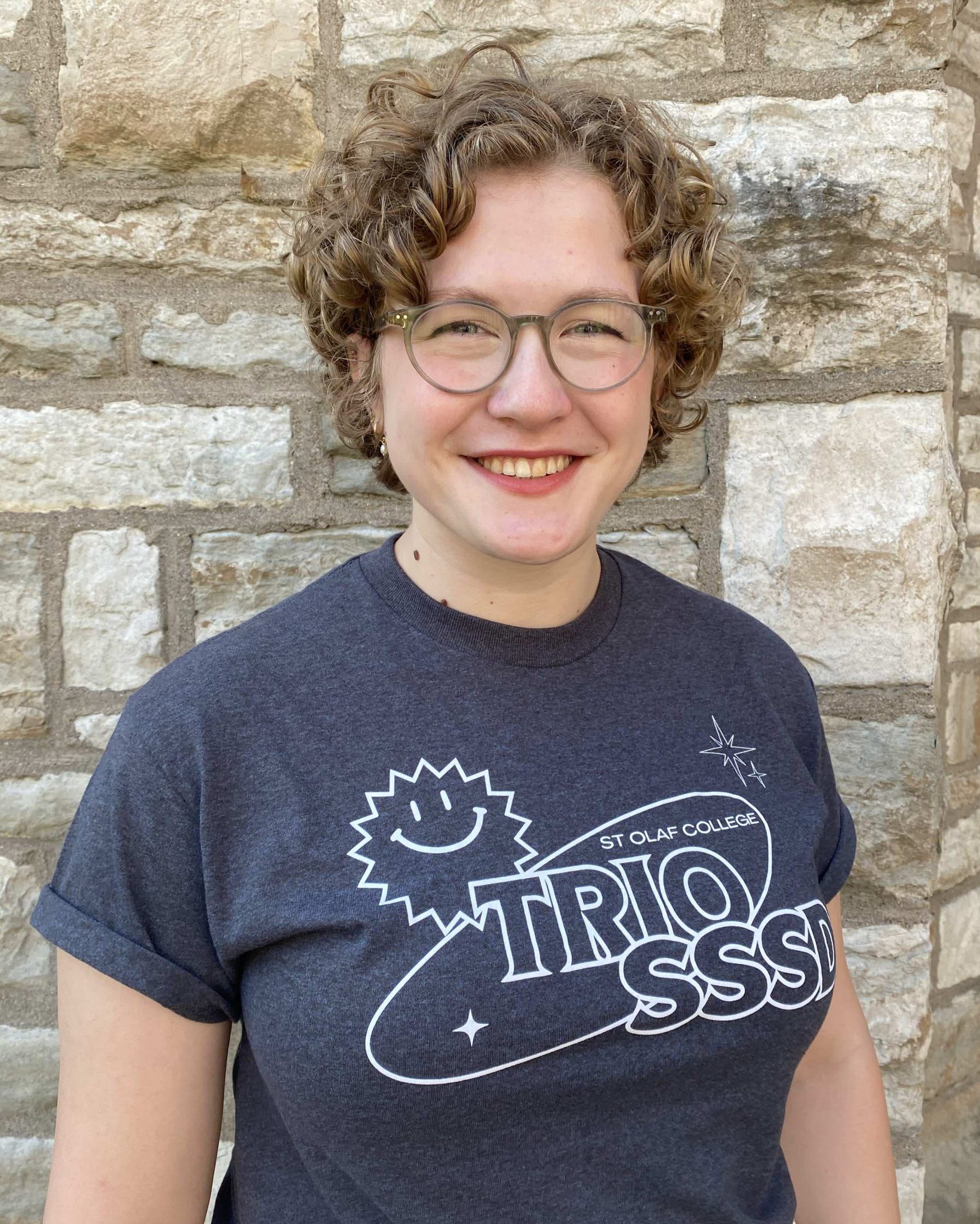 A smiling woman with glasses, short brownish-blonde hair and white skin. They are wearing the 2024 TRIO SSSD t-shirt and is standing in front of a limestone brick wall