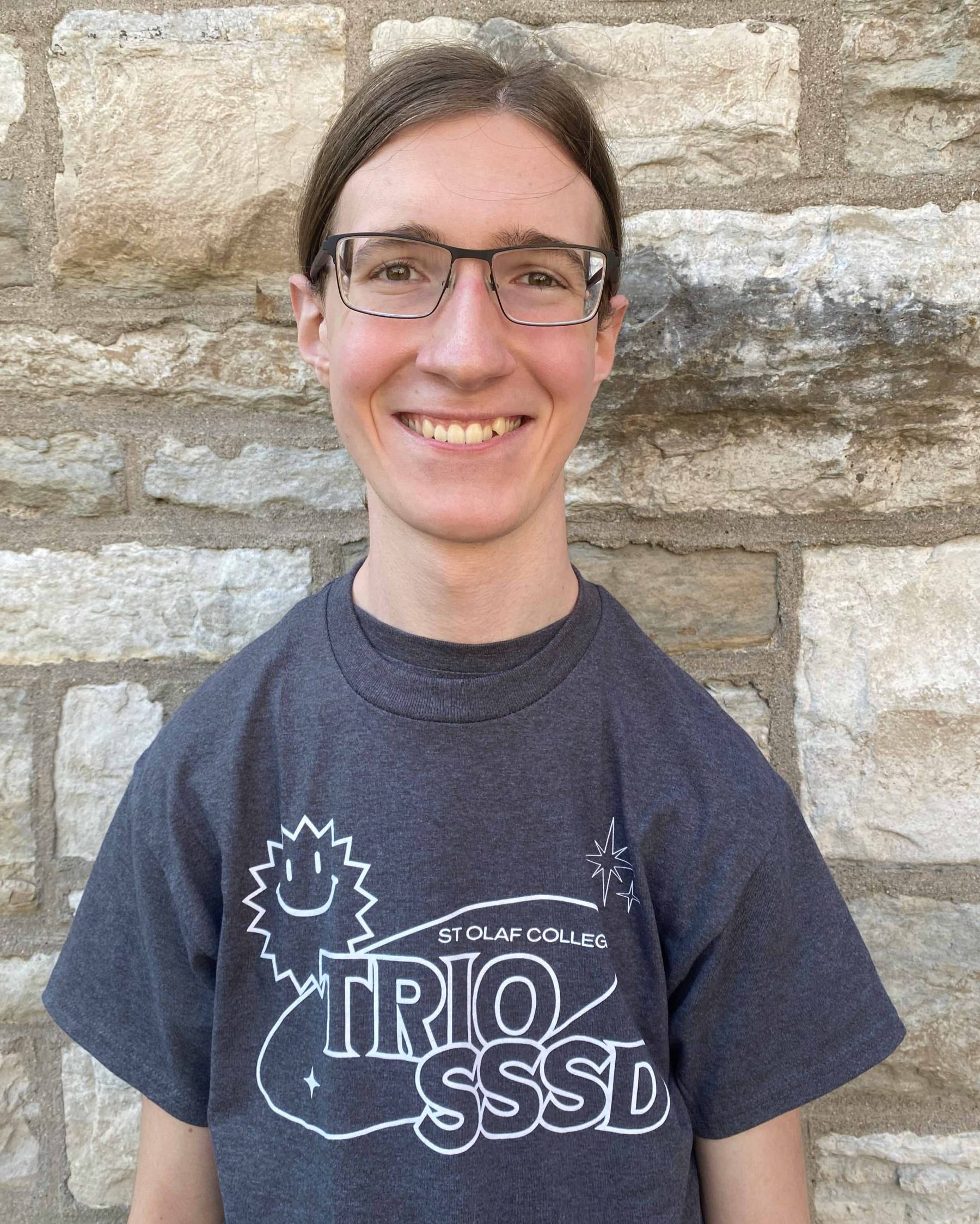 A smiling man with glasses, pulled back brown hair and white skin. They are wearing the 2024 TRIO SSSD t-shirt and standing in front of a limestone brick wall.