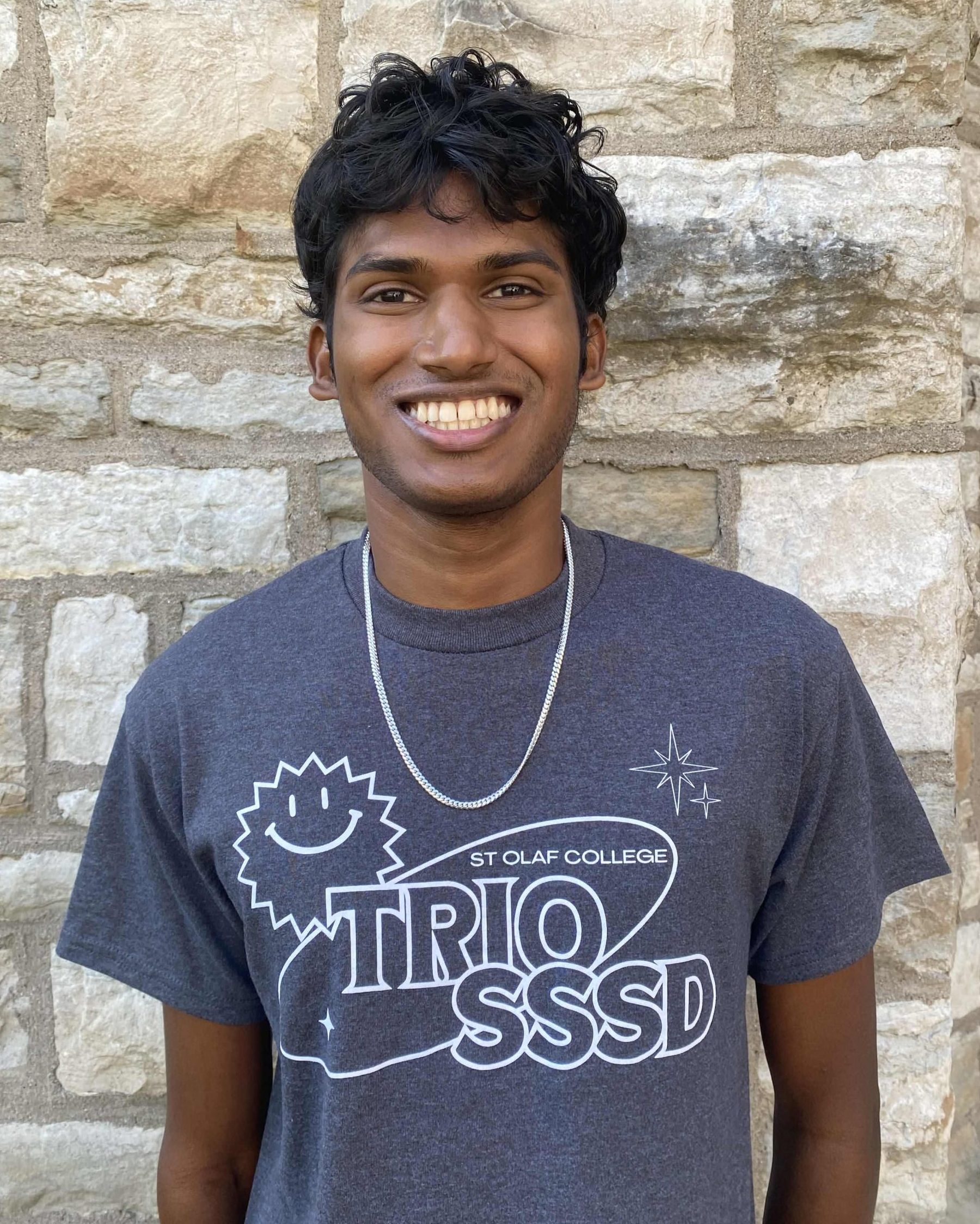 A smiling man with short black curly hair and dark brown skin. He is wearing the 2024 TRIO SSSD t-shirt and is standing in front of a limestone brick wall