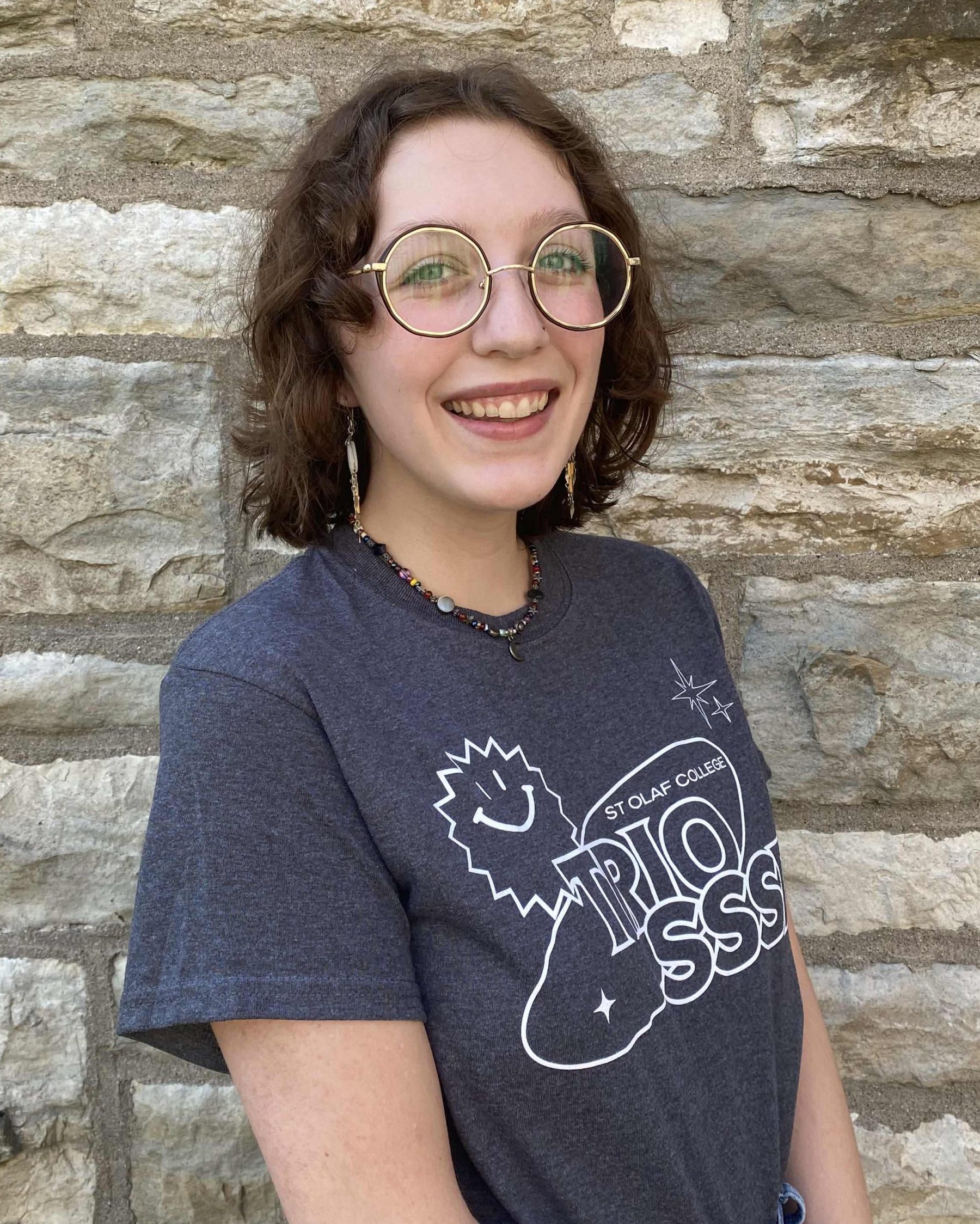 A smiling woman with glasses, medium-short curly brown hair and fair white skin. They are wearing the 2024 TRIO SSSD t-shirt and standing in front of a limestone brick wall.