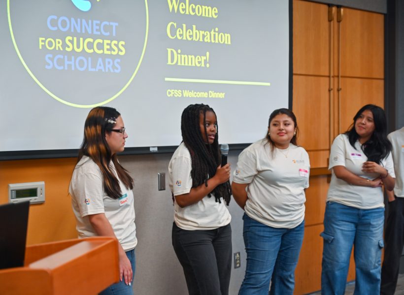 Group of 6 students waring light color t-shirts and jeans in front of a room. The screes shows a sign for Connect For Success Scholars. A student is holding a microphone.