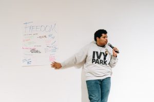 Black young adult male holding mic stands in front of a giant post-it titled "Freedom" with many remarks on it.