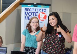 Two female young adults stand in front of "Register to Vote here" poster with smiles and thumbs up